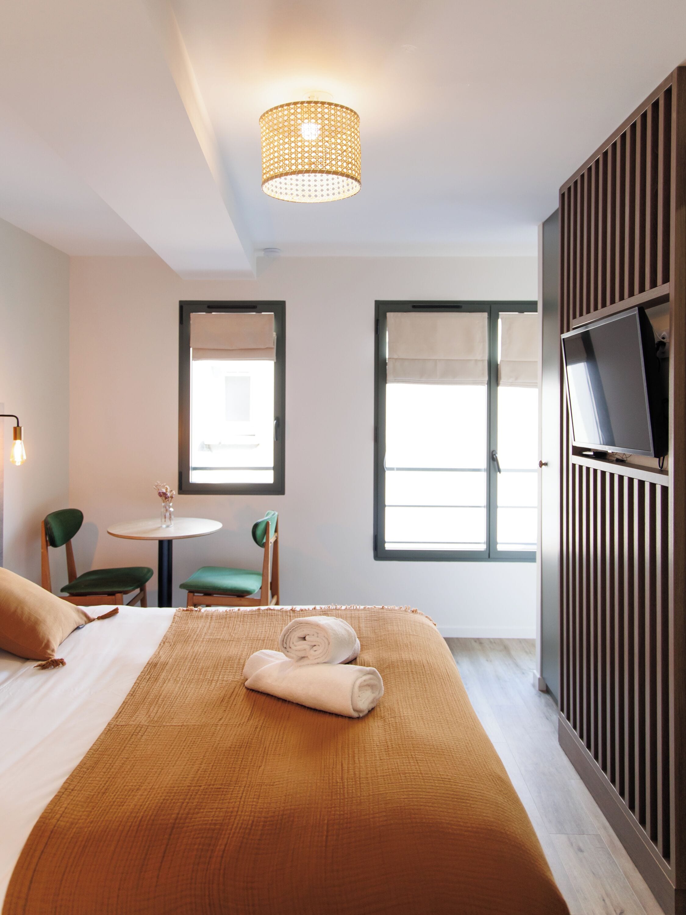 A hotel room with a bed, a small table with chairs, a television, and windows with curtains.