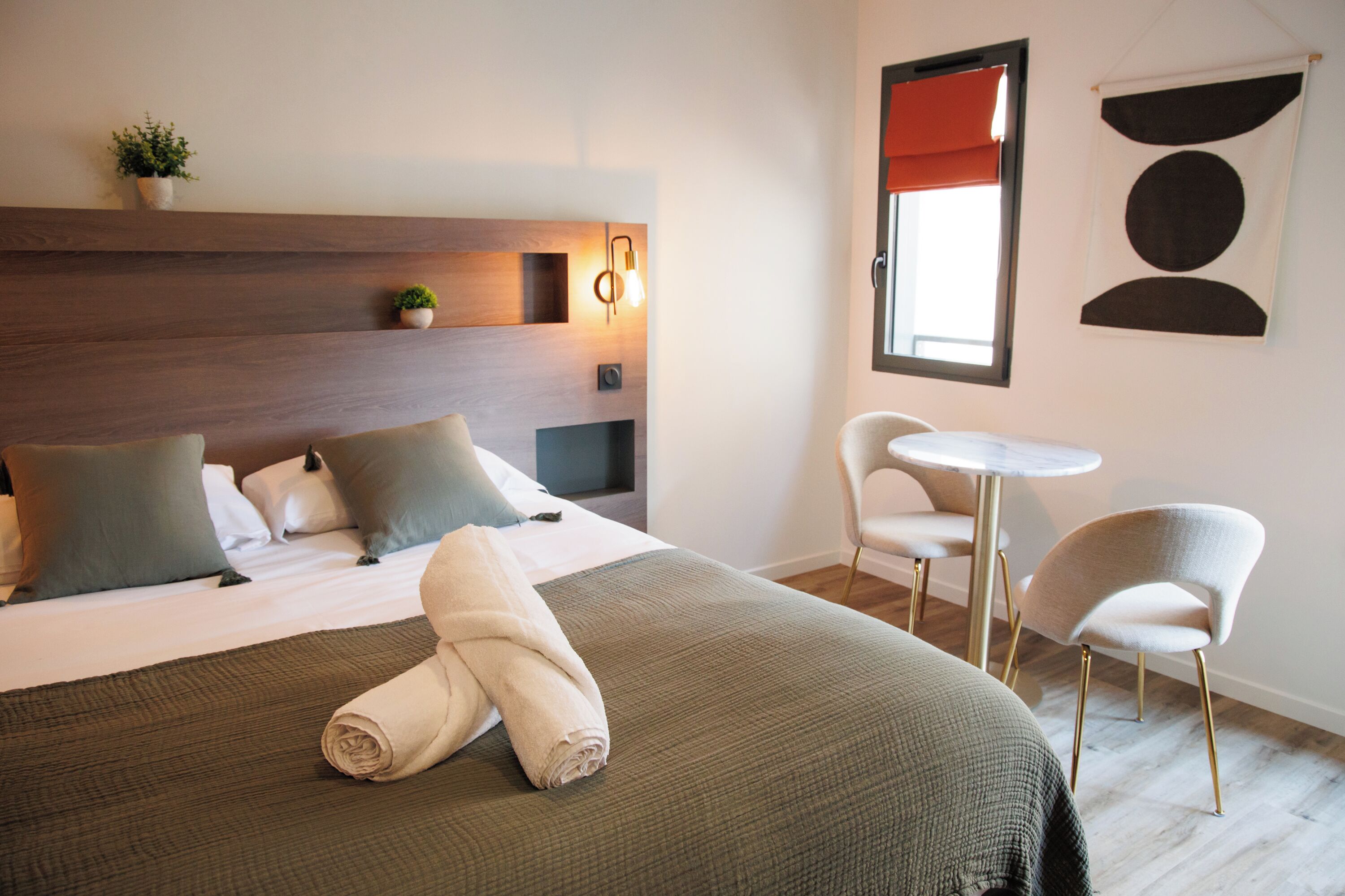 A hotel room with a bed, two chairs, a small table, a wall-mounted light, and a window with a red blind.