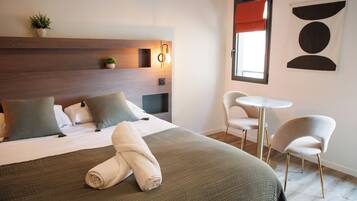 A hotel room with a bed, two chairs, a small table, a wall-mounted light, and a window with a red blind.