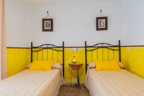 Quarto com duas camas, cada uma com um travesseiro amarelo, uma mesa pequena com abajur, e duas fotos emolduradas na parede.