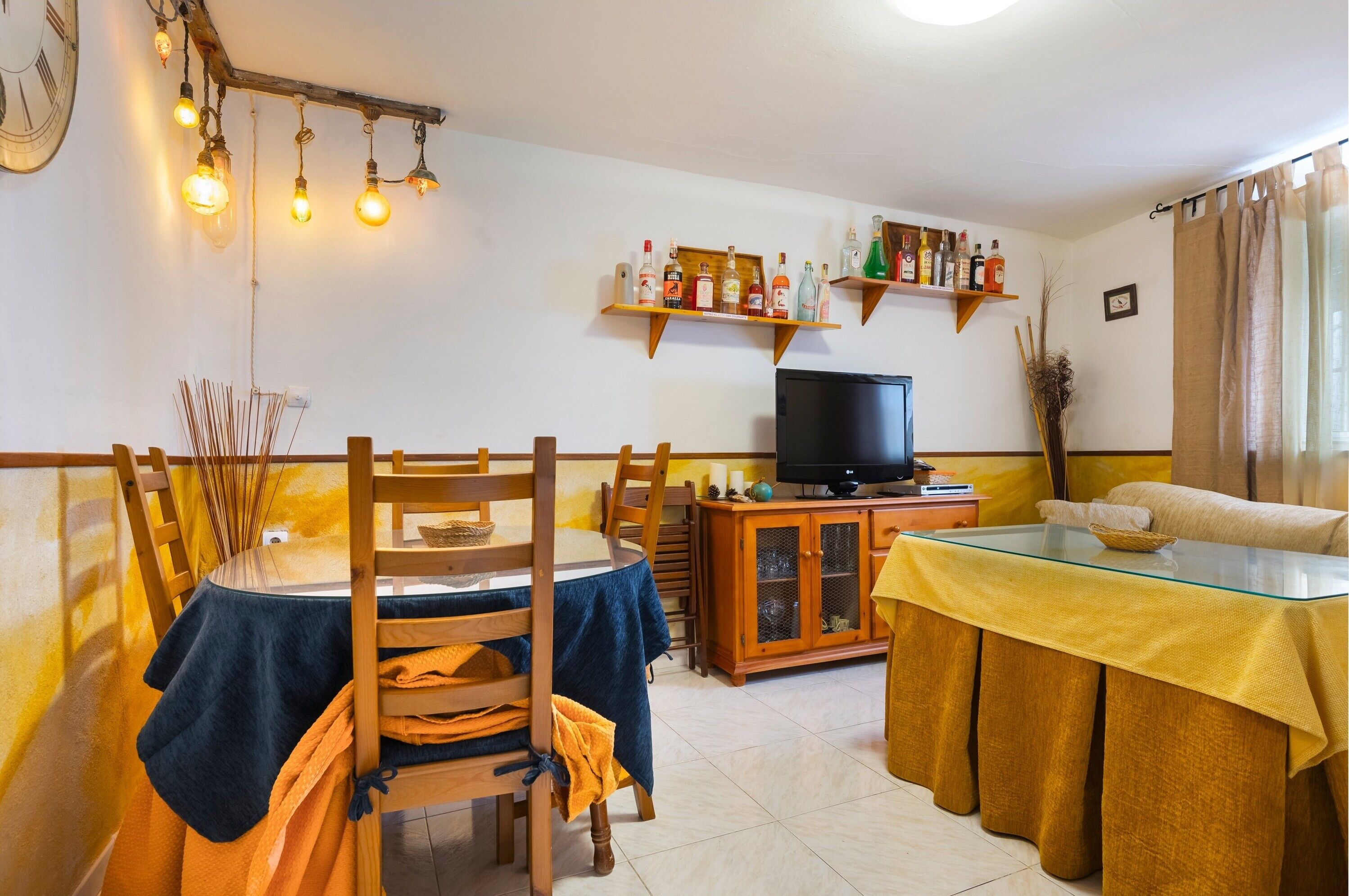 A dining area with a table, chairs, a television, and shelves with bottles.