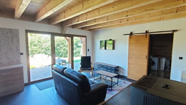 Un salon moderne avec un canapé en cuir noir, un plafond en bois, une porte-fenêtre coulissante donnant sur un patio et un poêle à bois encastré.