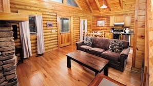 A log cabin interior with a wooden floor, a sofa, a coffee table, a stone wall, and a kitchen area.
