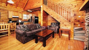 A log cabin interior with a wooden staircase, a living room with a sofa and coffee table, and a kitchen area.