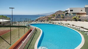 Una piscina curva con sillones de descanso, vista al mar y un edificio moderno al fondo.