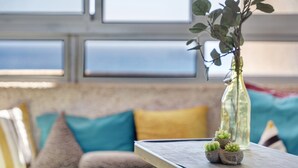 Vista despejada por una ventana al cielo, un sofá con cojines y una mesa con un florero y suculentas.
