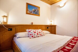 Un lit en bois avec une literie blanche, un coussin à motifs rouges et blancs, un tableau représentant un paysage de montagne et deux lampes fixées au mur.
