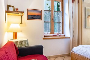 A room with a bed, a sofa, a lamp, a window with lace curtains, a picture on the wall, and a small table.