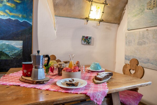 Une table en bois recouverte d’une nappe rose, une cafetière, un panier de fruits et une assiette avec un dessert.