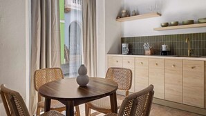 Un comedor con una mesa redonda de madera y sillas de mimbre, una cocina con armarios de madera y baldosas verdes, y una vista por una ventana con cortinas.