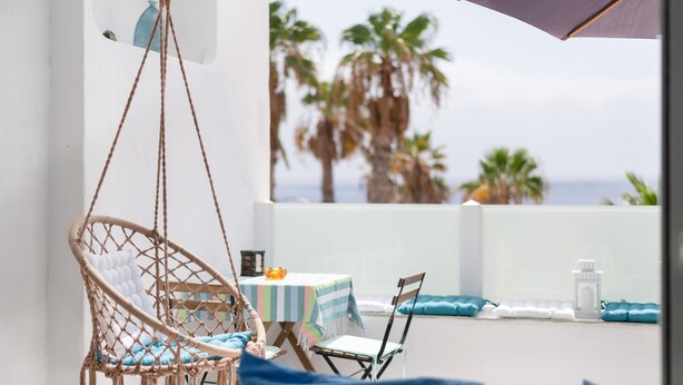 A balcony with a hanging chair, a small table with a striped tablecloth, and a view of palm trees.