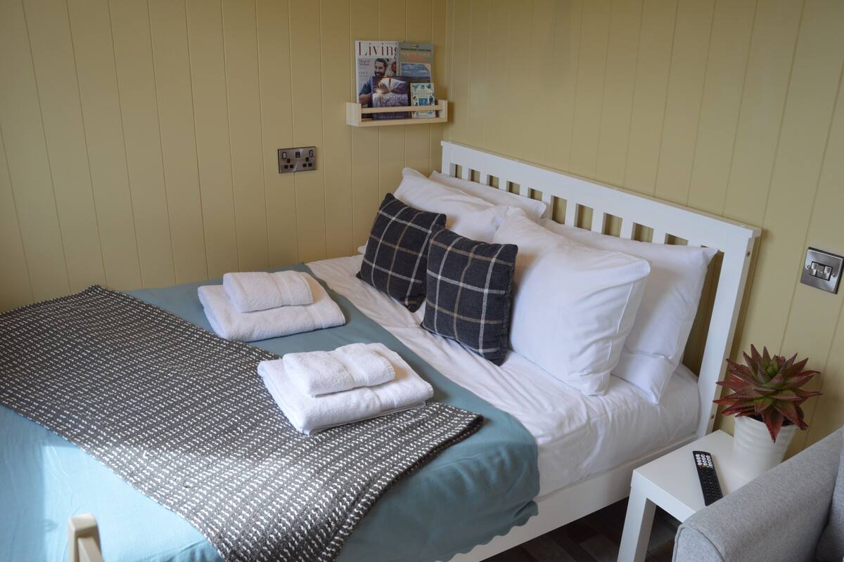 A neatly made bed with white linens, a blue bedspread, and a gray throw blanket. There is a white bedside table with a remote control and a potted plant.