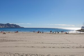 Ein weiter Sandstrand mit Menschen und Liegestühlen, ein klarer Himmel und ferne Berge.