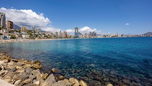 A coastal cityscape with clear blue water, rocky shoreline, and a variety of buildings including high-rise structures.