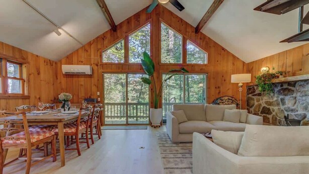 Una cabaña de madera espaciosa con comedor, sala de estar con chimenea de piedra y grandes ventanales con vistas al bosque.