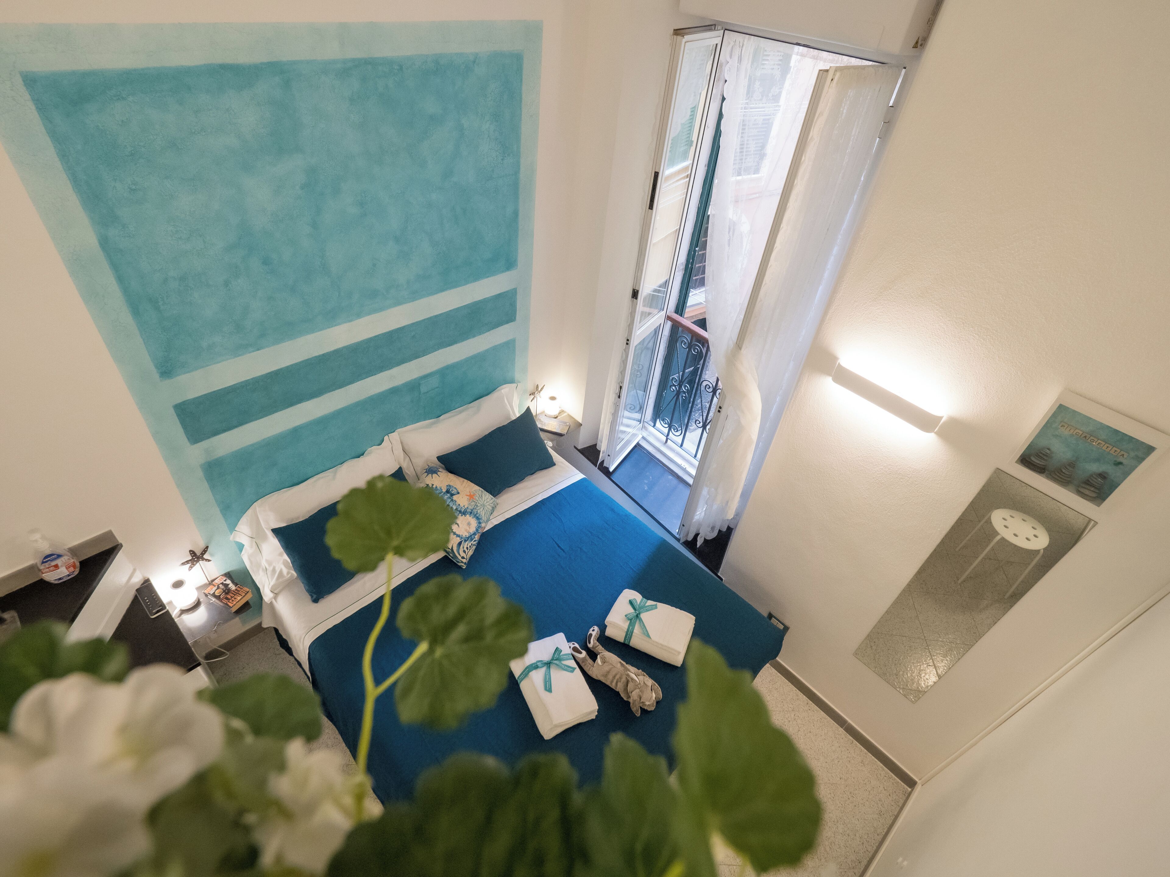 Habitación de hotel con un ventanal grande, una cama con ropa de cama azul, una mesita de noche con lámpara y un reloj en la pared.
