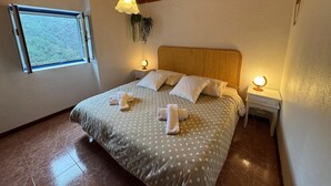 En snyggt bäddad säng med vita och grå prickiga sängkläder, två sänglampor och ett fönster med utsikt över träd.