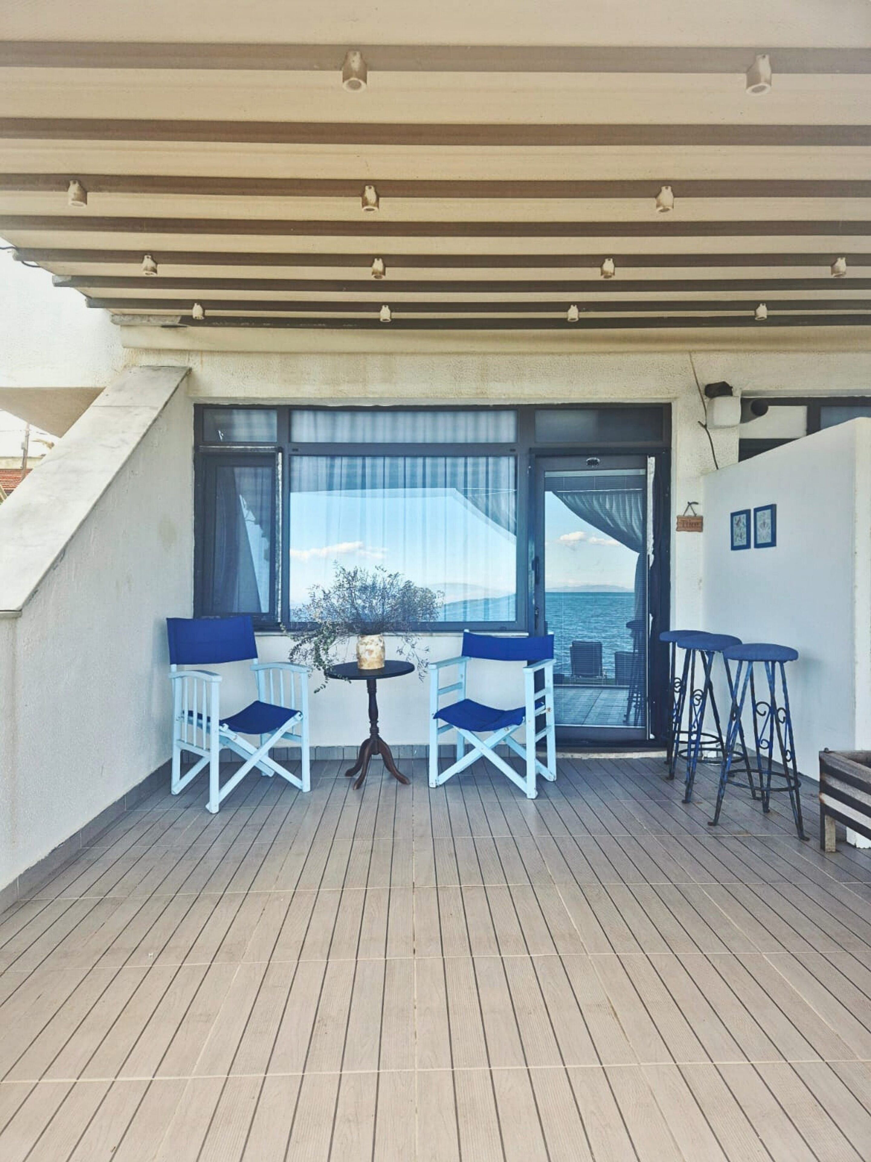 Un espace extérieur abrité avec deux chaises blanches et une table, une terrasse en bois et une vue sur la mer à travers de grandes fenêtres.