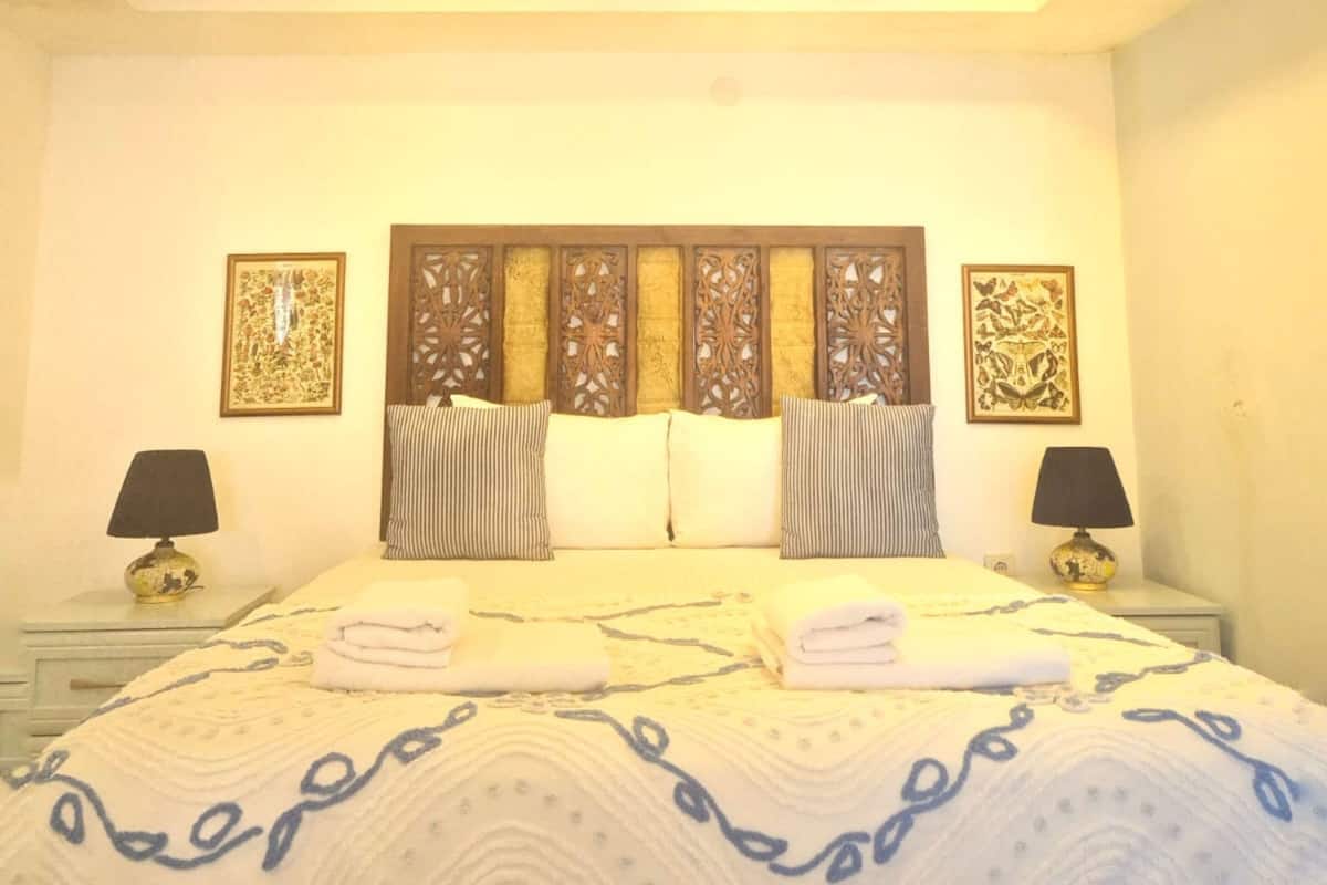 Quarto com cama grande, duas mesas de cabeceira, cabeceira decorativa e obra de arte na parede.