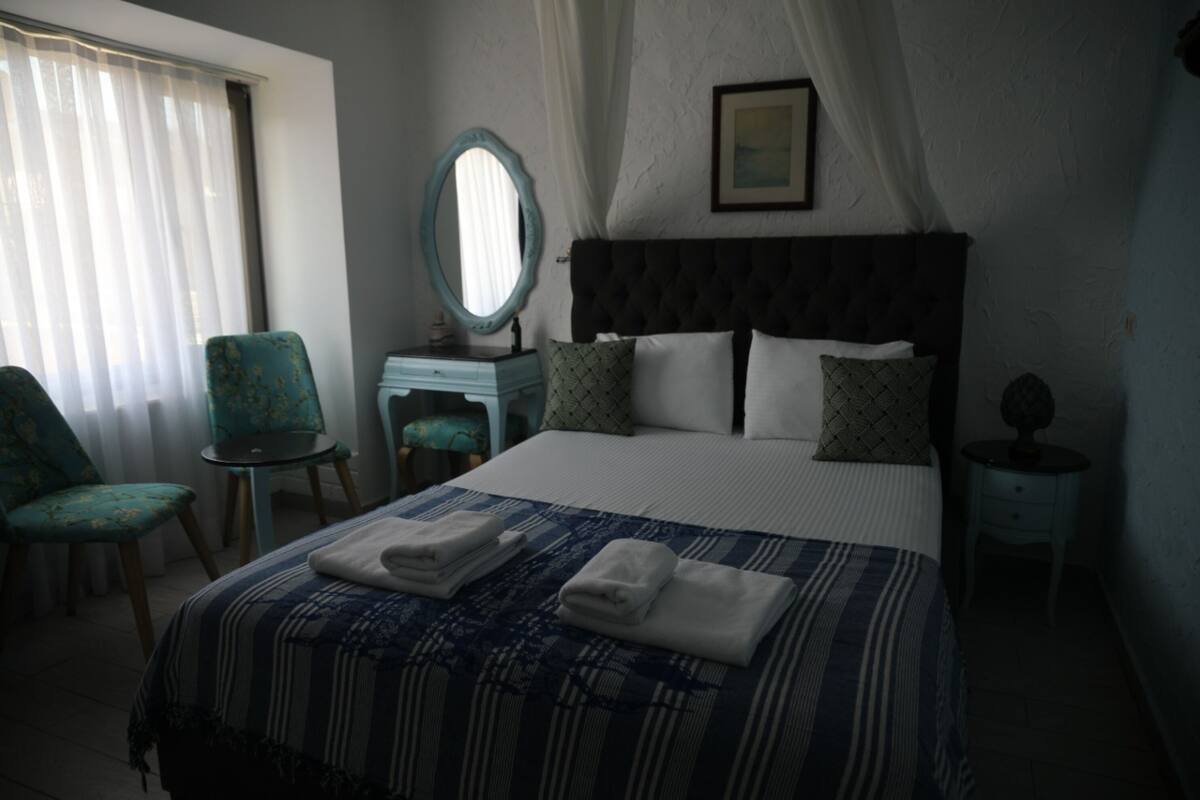 Quarto de hotel com cama, cadeira, mesa de cabeceira, espelho e janela com cortinas.
