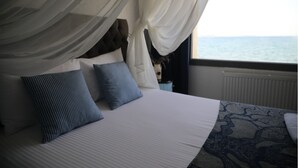Ein Bett mit Baldachin, zwei Kissen, ein gemustertes Teppich und Meerblick durch ein Fenster.