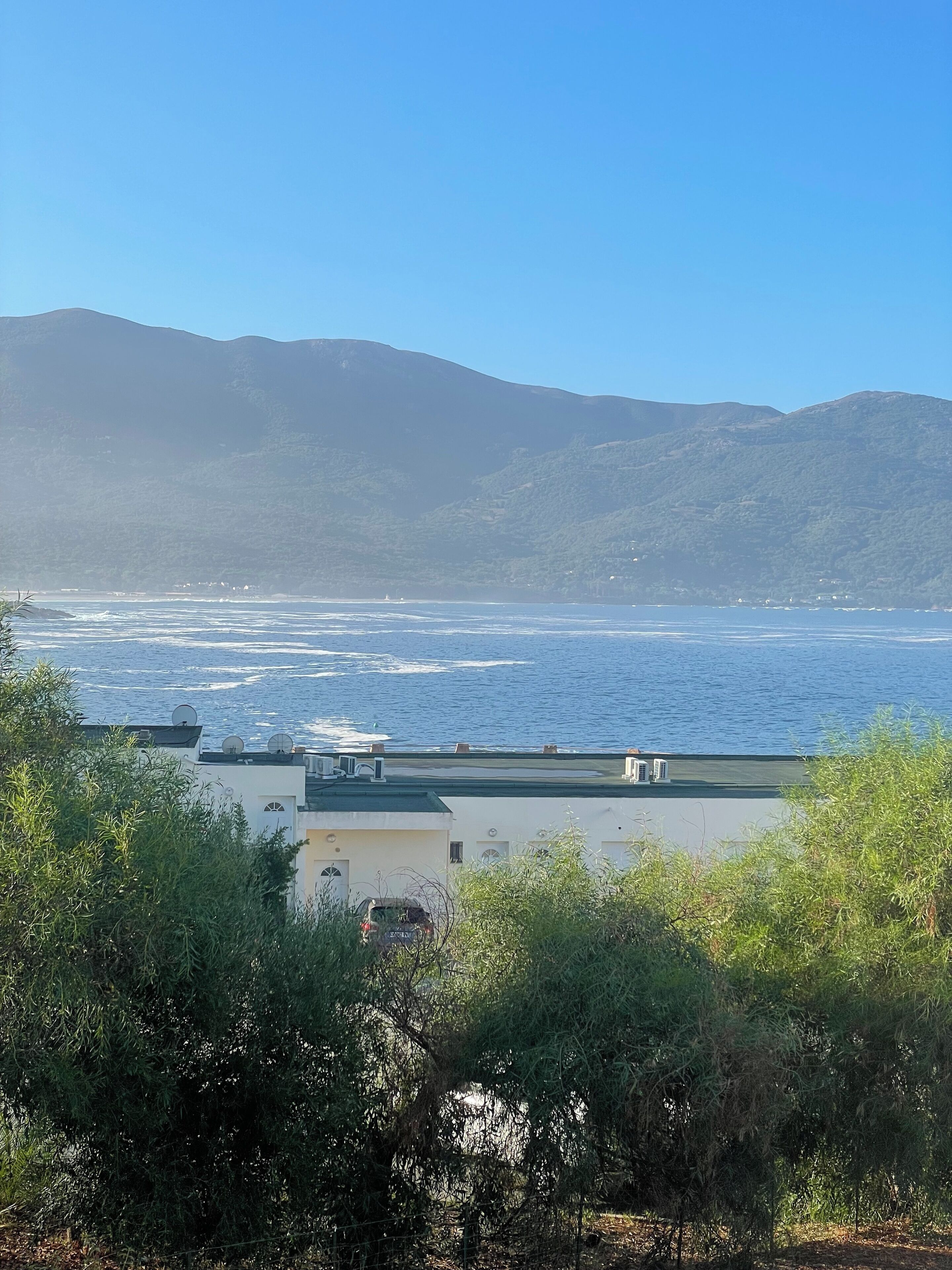 A coastal view with a building featuring satellite dishes and air conditioning units, surrounded by greenery.