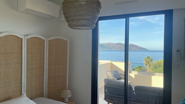 Ein Schlafzimmer mit Bett, Nachttisch, Lampe und Meerblick durch große Fenster.
