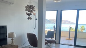 Ein modernes Wohnzimmer mit einer Couch, einem runden Couchtisch und einem Fernseher. Ein Sessel und eine Lampe sind ebenfalls vorhanden. Große Fenster bieten Blick auf das Meer und die Berge.