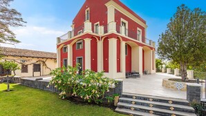 A red and cream multi-story building with a balcony, steps leading to the entrance, and a well-maintained lawn. - Badimà Villa with Pool (Aci Sant'Antonio)