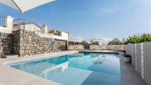 A swimming pool with stone walls, a white umbrella, and a view of a building with a balcony. - Badimà Villa with Pool (Aci Sant'Antonio)