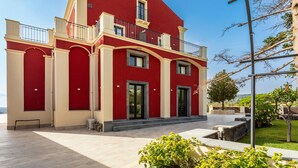 A red and beige building with a balcony, large windows, and a paved courtyard. - Badimà Villa with Pool (Aci Sant'Antonio)