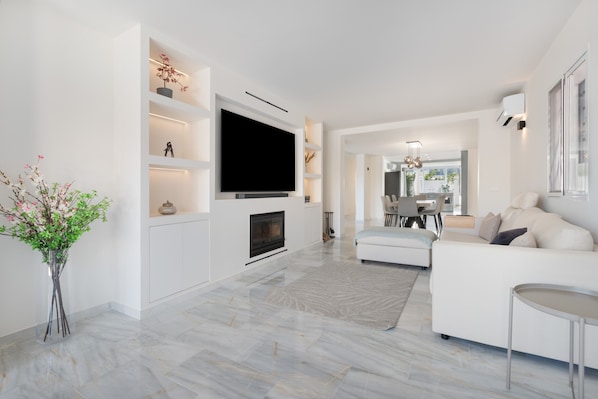 Un salon moderne avec un téléviseur à écran plat, une cheminée, un canapé blanc et un coin repas à l’arrière-plan.