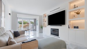 A modern living room with a sofa, a TV mounted on the wall, a fireplace, and a dining area with chairs.