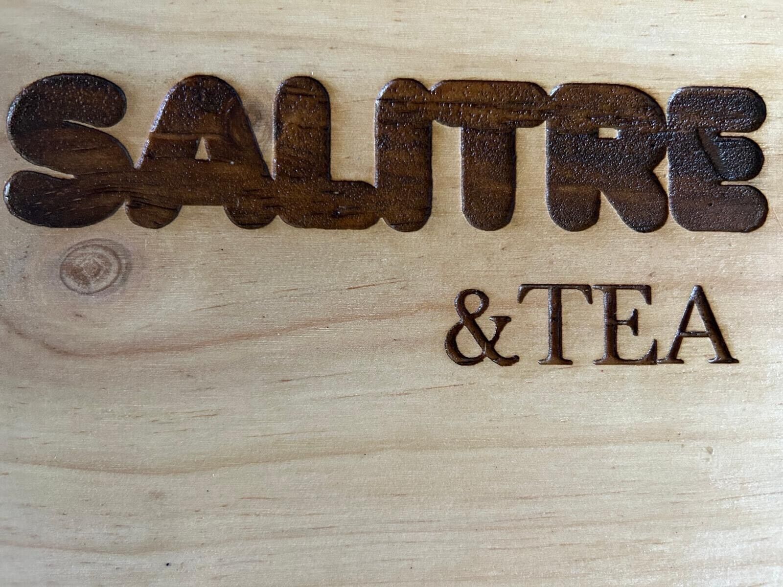 Une surface en bois sur laquelle sont gravés les mots « SALITRE & TEA » dans une police de caractères métallique et sombre.