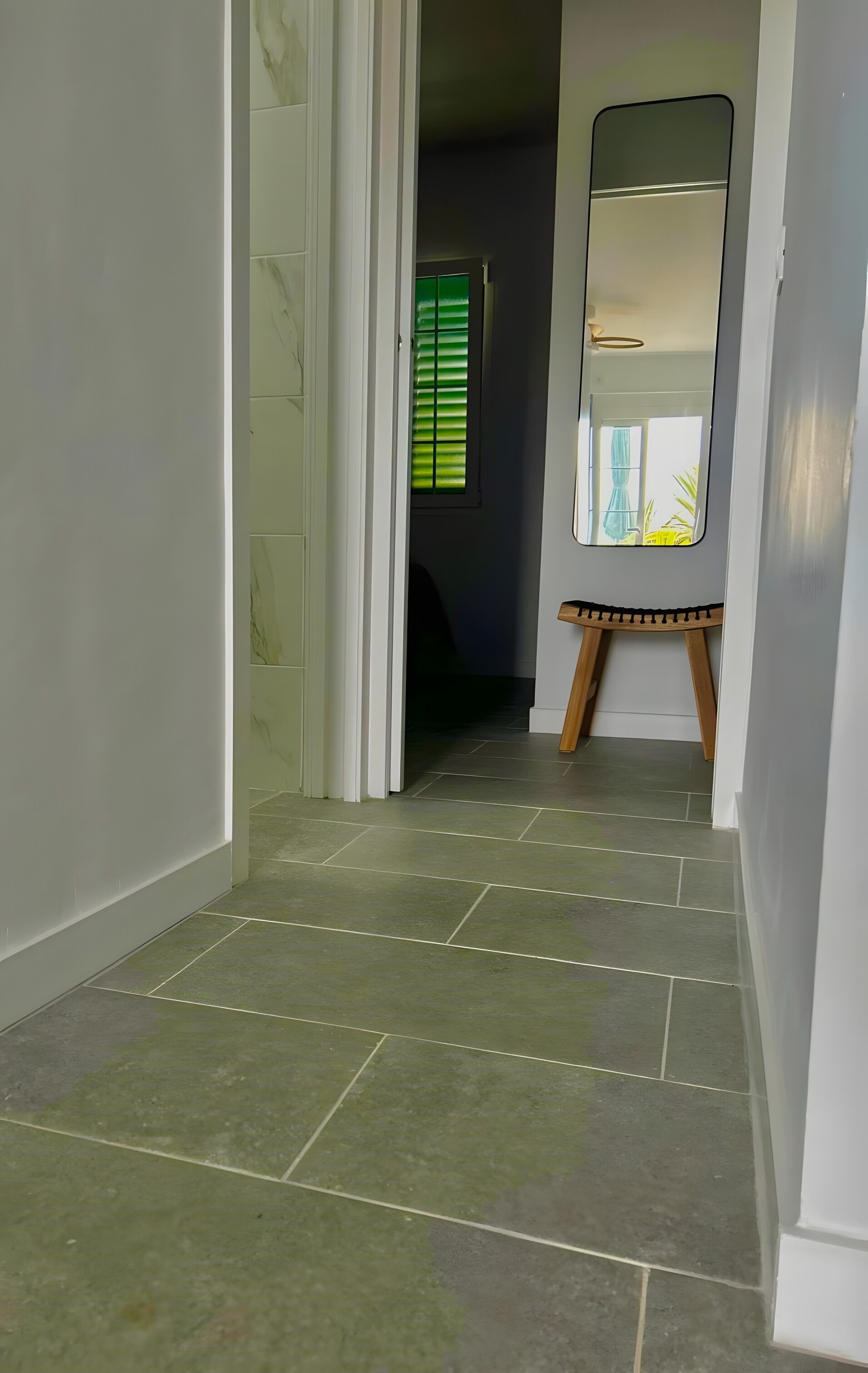 Un pasillo con suelo de baldosas, un espejo sobre un atril y una puerta que da a otra habitación.