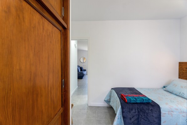 A bedroom with a bed, a wooden wardrobe, a bathroom with a mirror, and a hallway leading to another room.