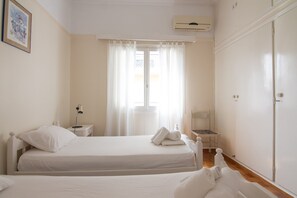 A bedroom with two beds, a bedside table with a lamp, a window with curtains, and an air conditioning unit. - Cozy Apartment Next to Alimos Beach (Alimos)