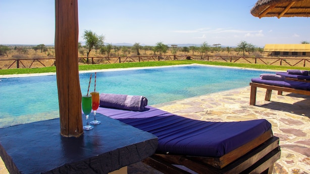 A poolside area with lounge chairs and a table, a clear view of the pool, and a thatched-roof structure in the background. - Africa Safari Serengeti Kusini  (Bariadi)