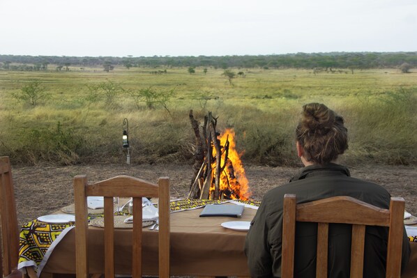 A person seated at a wooden table with a fire in the background, surrounded by a natural landscape. - Africa Safari Serengeti Kusini (Bariadi)
