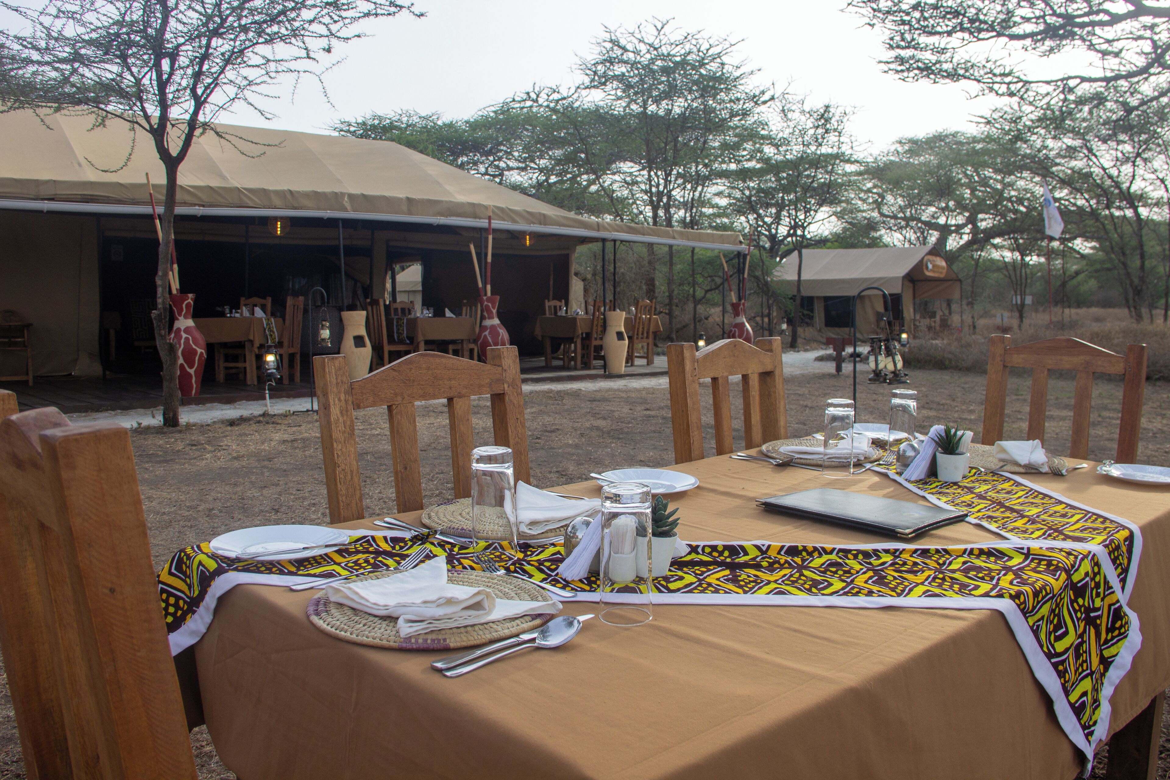 An outdoor dining area with tables set for a meal, surrounded by wooden chairs and a tent structure in the background.