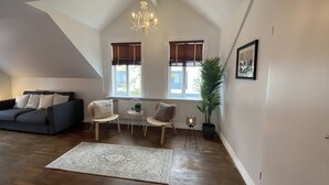 Ein großzügiger Loft mit hohen Decken, einem dunkelgrauen Sofa, zwei Sesseln, einem kleinen Tisch, einer Topfpflanze und einer Kristallleuchter.