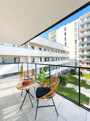 A balcony with two wicker chairs and a table, a potted plant, and a view of a modern building complex. - Lumina 3-room apart on Drewnowska (Lódz)