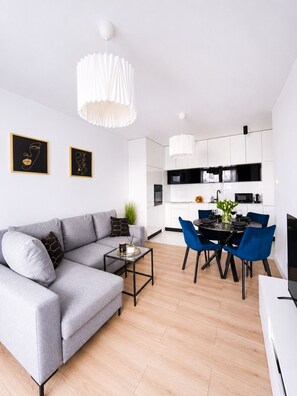 A modern living room with a grey sofa, a coffee table, and a dining area with blue chairs. - Lumina 3-room apart on Drewnowska (Lódz)