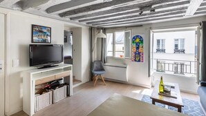 A living room with a television, a chair, a table with a bottle, and a window with a balcony view.