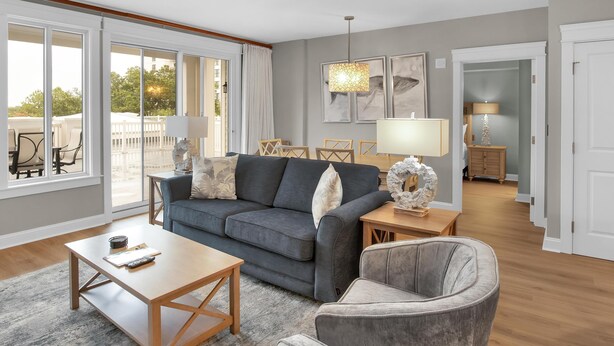 A spacious living room with a grey sofa, a wooden coffee table, and a grey armchair. There is a ceiling fan, a dining area with a table and chairs, and a hallway leading to another room.