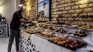 A buffet with various food items, a person serving, and a brick wall with a painting. - HOTEL LA PERLE DU SUD (Ouarzazate)