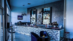 A modern bar area with a central bar counter, multiple seating options including bar stools and chairs, and a decorative wall with shelves displaying bottles. - HOTEL LA PERLE DU SUD (Ouarzazate)