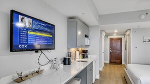 A modern hotel room with a kitchen area, a flat-screen TV mounted on the wall, and a wooden shelf with decorative items.