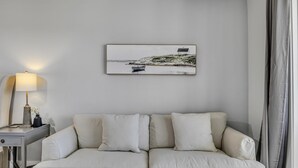 A modern living room with a sofa, a side table, a lamp, and a framed picture of a coastal scene.