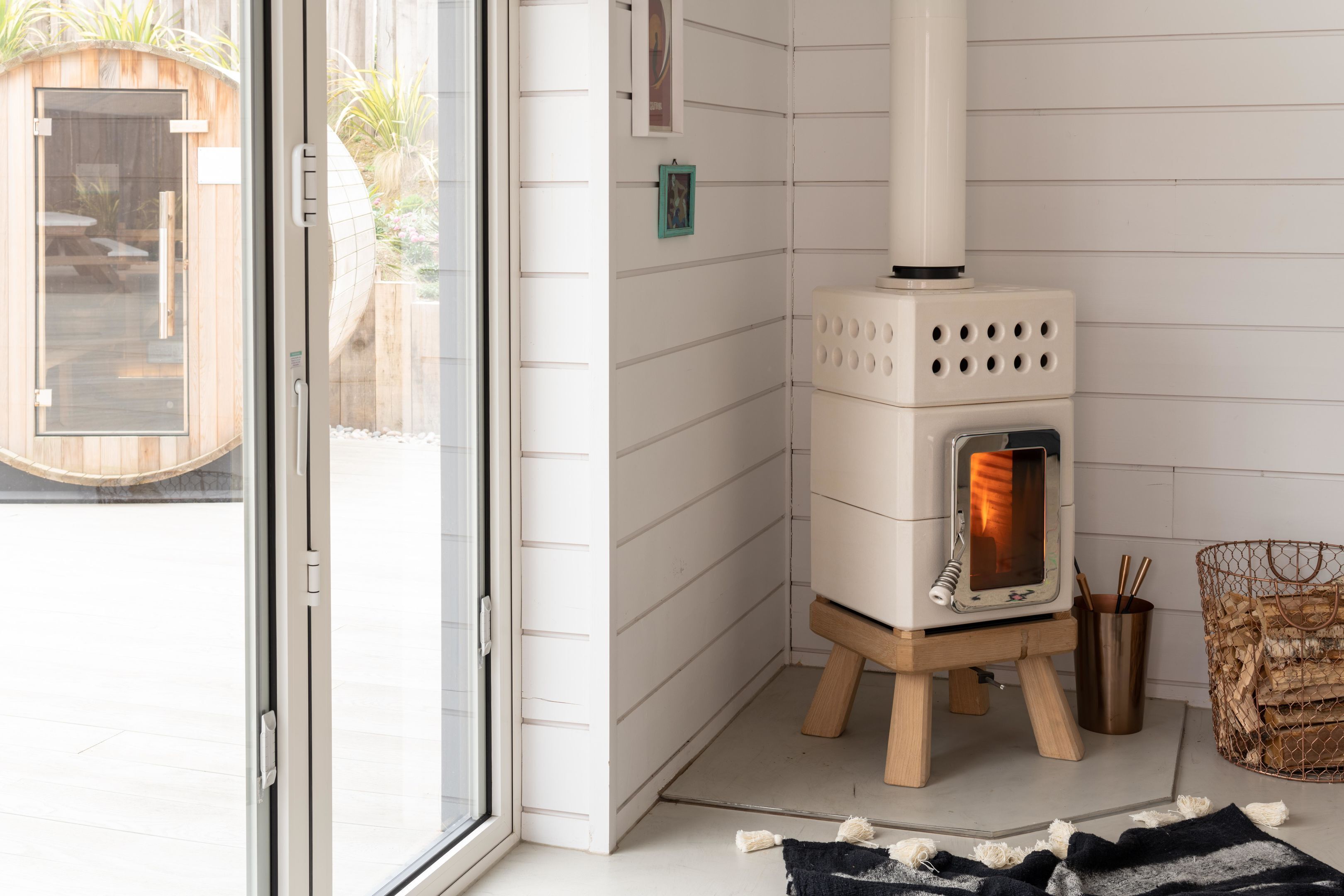 A modern, compact wood-burning stove in a corner of a room with white paneled walls and a glass door leading to an outdoor area.